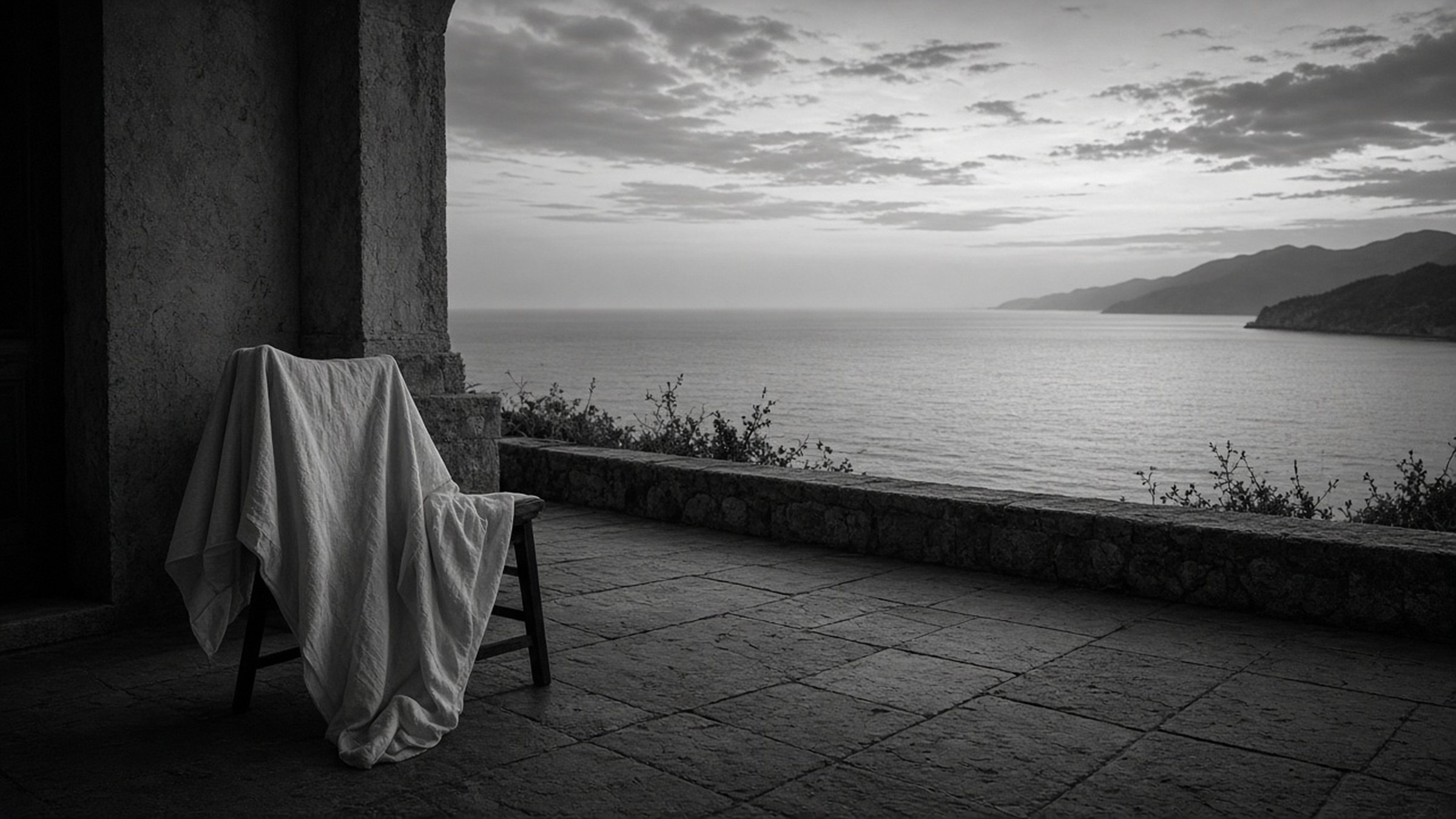 A tranquil black-and-white terrace of weathered stone overlooking a calm sea, with a chair draped in white linen